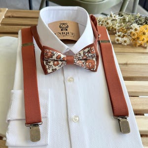 Peut inclure: Une chemise blanche à motif texturé, assortie d'un nœud papillon et de bretelles couleur rouille. Le nœud papillon présente un motif floral aux tons marron et crème. Les bretelles ont des attaches métalliques. L'étiquette indique "UNIQUE Accessories".