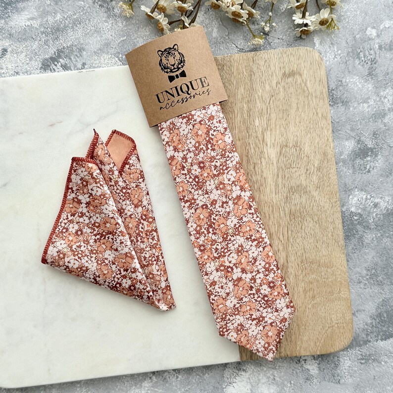 K&ouml;nnte beinhalten: Ein Set aus Krawatte und Einstecktuch mit floralem Muster. Die Krawatte ist in einer braunen Papph&uuml;lle mit dem Text "UNIQUE accessories" und einem Tiger-Logo verpackt. Das Einstecktuch ist gefaltet und hat ein passendes Blumenmuster. Das Muster umfasst Braunt&ouml;ne, Pfirsich und Wei&szlig;.