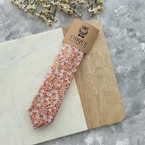 K&ouml;nnte beinhalten: Eine Krawatte mit Pfirsich- und wei&szlig;en Blumenmustern, pr&auml;sentiert auf einem Holzbrett. Die Krawatte ist mit einem braunen Etikett mit der Aufschrift "UNIQUE accessories" und einem Tigerlogo versehen. Das Design der Krawatte ist ein sich wiederholendes Blumenmuster.