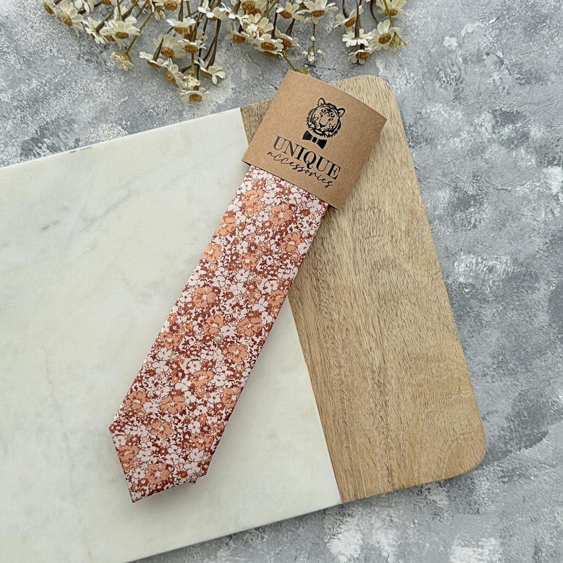 K&ouml;nnte beinhalten: Eine Krawatte mit Pfirsich- und wei&szlig;en Blumenmustern, pr&auml;sentiert auf einem Holzbrett. Die Krawatte ist mit einem braunen Etikett mit der Aufschrift "UNIQUE accessories" und einem Tigerlogo versehen. Das Design der Krawatte ist ein sich wiederholendes Blumenmuster.