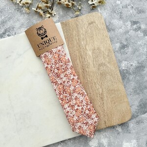 K&ouml;nnte beinhalten: Eine Krawatte mit Pfirsich- und wei&szlig;en Blumenmustern, pr&auml;sentiert mit ihrer Markenverpackung. Die Verpackung zeigt ein Tigerlogo und die Worte "UNIQUE accessories". Die Krawatte wird auf einer Marmor- und Holzoberfl&auml;che pr&auml;sentiert.