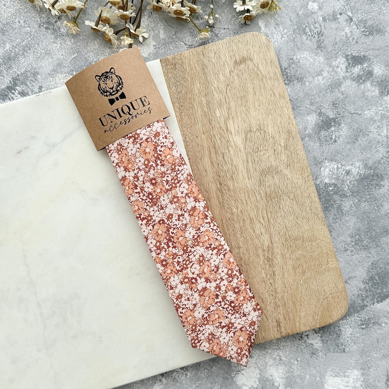 K&ouml;nnte beinhalten: Eine Krawatte mit Pfirsich- und wei&szlig;en Blumenmustern, pr&auml;sentiert mit ihrer Markenverpackung. Die Verpackung zeigt ein Tigerlogo und die Worte "UNIQUE accessories". Die Krawatte wird auf einer Marmor- und Holzoberfl&auml;che pr&auml;sentiert.