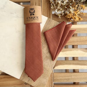 May include: A rust-colored tie and matching pocket square set. The tie is displayed with a branded tag that reads "UNIQUE ACCESSORIES" and features a tiger logo. The pocket square has a red border. The set is presented on a wooden surface.