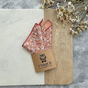 K&ouml;nnte beinhalten: Ein Einstecktuch mit Pfirsich- und wei&szlig;en Blumenmustern und einem rot gen&auml;hten Rand. Es wird auf einer Marmor- und Holzoberfl&auml;che pr&auml;sentiert, zusammen mit einem braunen Etikett mit dem Text "UNIQUE accessories".