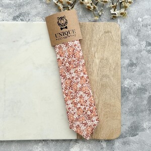 K&ouml;nnte beinhalten: Eine Krawatte mit floralem Muster in Pfirsich-, Wei&szlig;- und Braunt&ouml;nen, pr&auml;sentiert auf einem Holzbrett. Die Krawatte ist mit einem braunen Pappanh&auml;nger mit einem Tigerlogo und den Worten "UNIQUE accessories" verpackt.