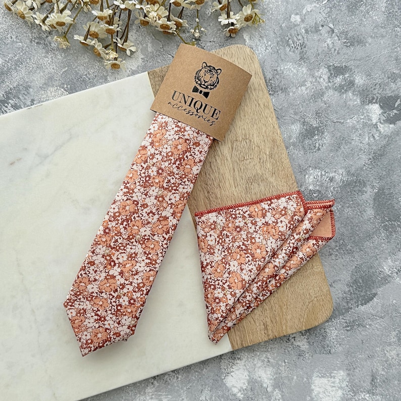 K&ouml;nnte beinhalten: Ein Set aus Krawatte und Einstecktuch mit floralem Muster. Krawatte und Einstecktuch weisen ein sich wiederholendes Muster aus kleinen, pfirsichfarbenen Blumen auf braunem Hintergrund auf. Das Set ist mit einem braunen Etikett mit der Aufschrift "UNIQUE accessories" verpackt.