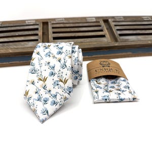 May include: A white tie with a blue and brown floral pattern. The tie is paired with a matching pocket square. The pocket square is folded and placed on a brown paper tag with the text "UNIQUE accessories".