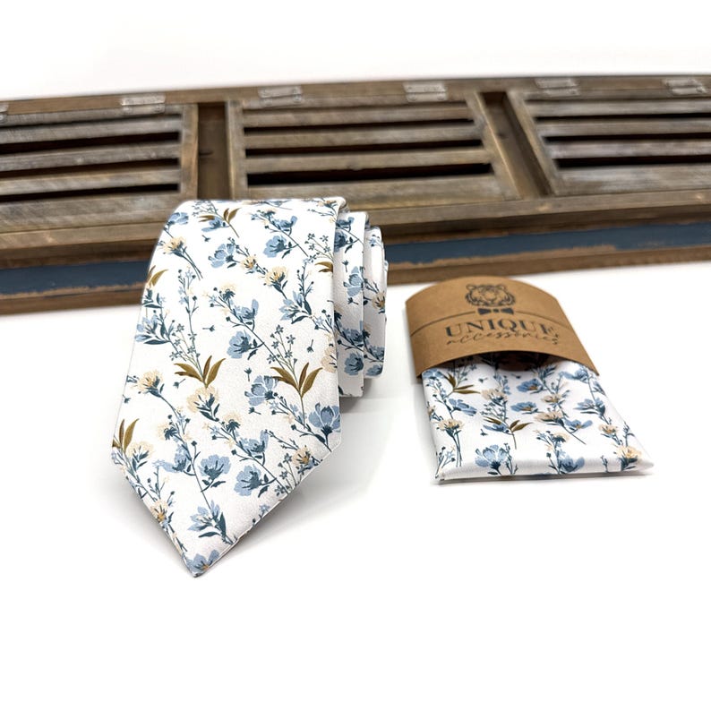 May include: A white tie with a blue and brown floral pattern. The tie is paired with a matching pocket square. The pocket square is folded and placed on a brown paper tag with the text "UNIQUE accessories".