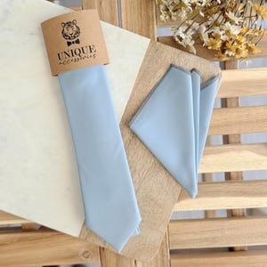 May include: A light blue tie and pocket square set. The tie is displayed with its original packaging, which features a tiger logo and the words "UNIQUE accessories". The pocket square has a gray stitched border.