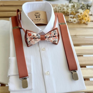 Può includere: Una camicia bianca con un papillon floreale e bretelle color ruggine. Il papillon presenta un motivo floreale con accenti arancioni, bianchi e marroni. Le bretelle hanno clip metalliche. Un'etichetta con il testo "UNIQUE accessories" è visibile.