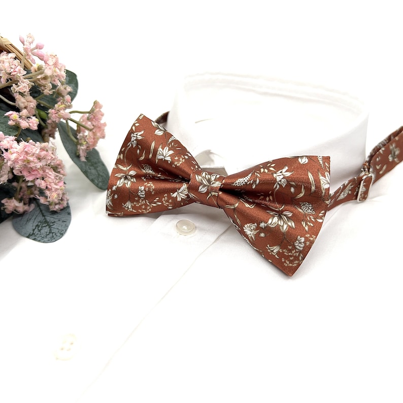 Peut inclure: Un n&oelig;ud papillon marron avec un motif floral blanc. Le n&oelig;ud papillon est en tissu soyeux et a un design classique.
