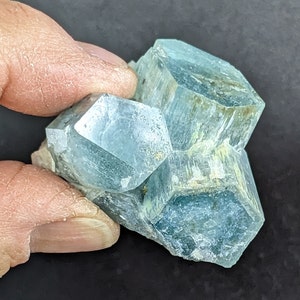 以下が含まれることがあります： 薄い青色のアクアマリン結晶のクラスター。結晶は六角形で半透明です。色は淡い青から中程度の青まで変化し、一部の領域は緑色を示しています。結晶は手で持たれています。