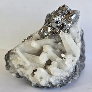 Può includere: Un campione di minerale naturale con un gruppo di cristalli di quarzo trasparente e un minerale metallico color argento. I cristalli di quarzo sono appuntiti e raggruppati, su una matrice grigio scuro. Il minerale metallico ha una superficie riflettente e lucida.