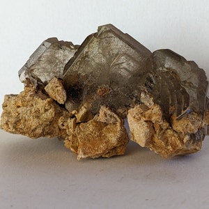 Può includere: Un gruppo di cristalli di quarzo fumé con una struttura geometrica a strati. I cristalli sono di un colore grigio-brunastro traslucido, posti su una base di roccia ruvida color beige. La composizione è naturale e terrosa.