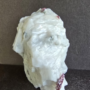 May include: A large, rough white mineral specimen with a crystalline structure. Small, dark red crystals are embedded in the white stone. The mineral has a textured surface and irregular shape, with a dark background.