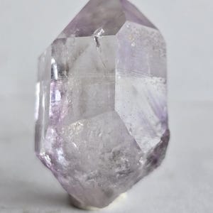 May include: A translucent amethyst crystal with a faceted, geometric shape. The crystal has a pale purple hue with areas of clear transparency. The crystal is standing upright, showcasing its natural form and texture.