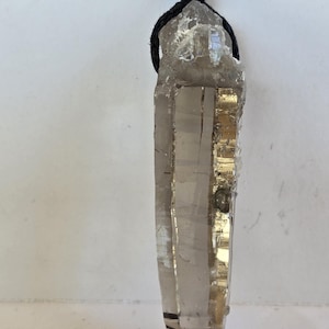 May include: A clear quartz crystal pendant with a black cord. The crystal is a light brown colour with a pointed tip and a rough, natural texture. The cord is knotted at the top for wearing.