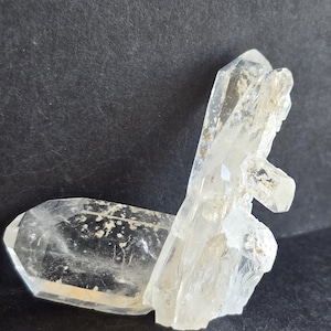 Puede incluir: Un grupo de cristales de cuarzo transparente con una punta principal horizontal unida a una formación vertical. El espécimen mineral muestra facetas naturales, superficies translúcidas y pequeñas inclusiones internas.