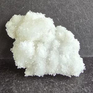 May include: A cluster of small, clear, crystalline structures, possibly quartz or a similar mineral, with a slightly frosted appearance. The crystals are densely packed, creating a textured, irregular shape. The background is a dark gray surface.