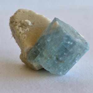 以下が含まれることがあります： 長方形の形をした水色ののアクアマリンの結晶が、ベージュ色のざらざらした質感の石に付いています。アクアマリンは半透明で、ベージュ色の石は多孔質に見えます。白い背景に配置されています。