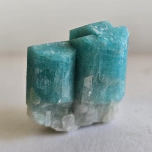 Puede incluir: Un grupo de cristales de amazonita, que muestra un vibrante tono turquesa con una estructura cristalina. La base es blanca contrastante, proporcionando una estética natural y terrosa. La superficie de la piedra tiene una apariencia sutil y texturizada.