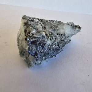 May include: A mineral specimen with a mix of grey, black, and white colours. The rock has a crystalline structure with visible metallic flecks. The overall shape is irregular, with a jagged edge.