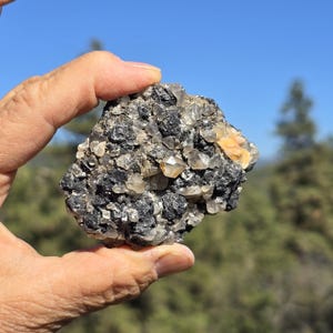 May include: A raw mineral specimen featuring a dark, textured matrix embedded with numerous small, translucent, multifaceted quartz crystals and a single prominent peach-colored cluster, showcasing natural geological formations and varied textures.