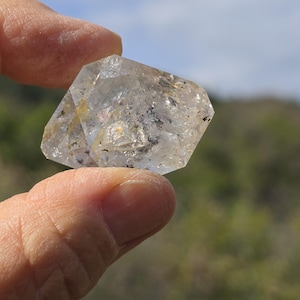 Puede incluir: Un cristal de cuarzo transparente y multifacético con fracturas internas visibles e inclusiones minerales oscuras. La piedra tiene una forma geométrica angular con bordes afilados y una superficie pulida y reflectante que capta la luz.