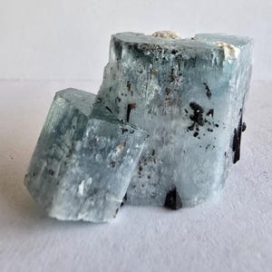 以下が含まれることがあります： アクアマリンの結晶の塊で、暗いインクルージョンを伴う淡い青色を示しています。結晶は白い背景に対して粗く自然な質感を持っています。構成は、1つ大きく、1つ小さい2つの主要な結晶形成を特徴としています。