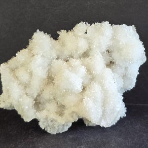May include: A cluster of small, clear quartz crystals. The crystals have a sparkling, frosted appearance and are densely packed together. The overall color is a pale, translucent white, with a slightly rough texture. The quartz formation sits on a dark surface.