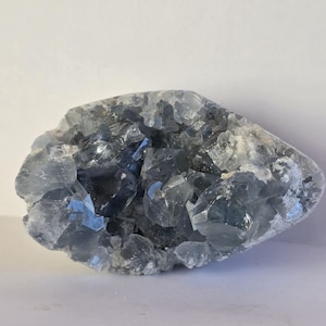 May include: A celestite geode featuring a cluster of translucent, pale blue, angular crystals. The mineral specimen has a jagged, irregular shape with varying depths and facets that reflect light across its crystalline surface.