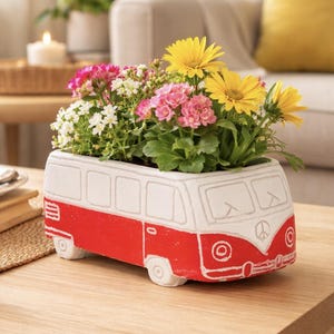 Puede incluir: Una maceta de cerámica con forma de furgoneta vintage, pintada en rojo y blanco con un símbolo de la paz en la parte delantera. La maceta está llena de una variedad de flores en flor, incluyendo margaritas amarillas, kalanchoe rosa y pequeñas flores blancas.