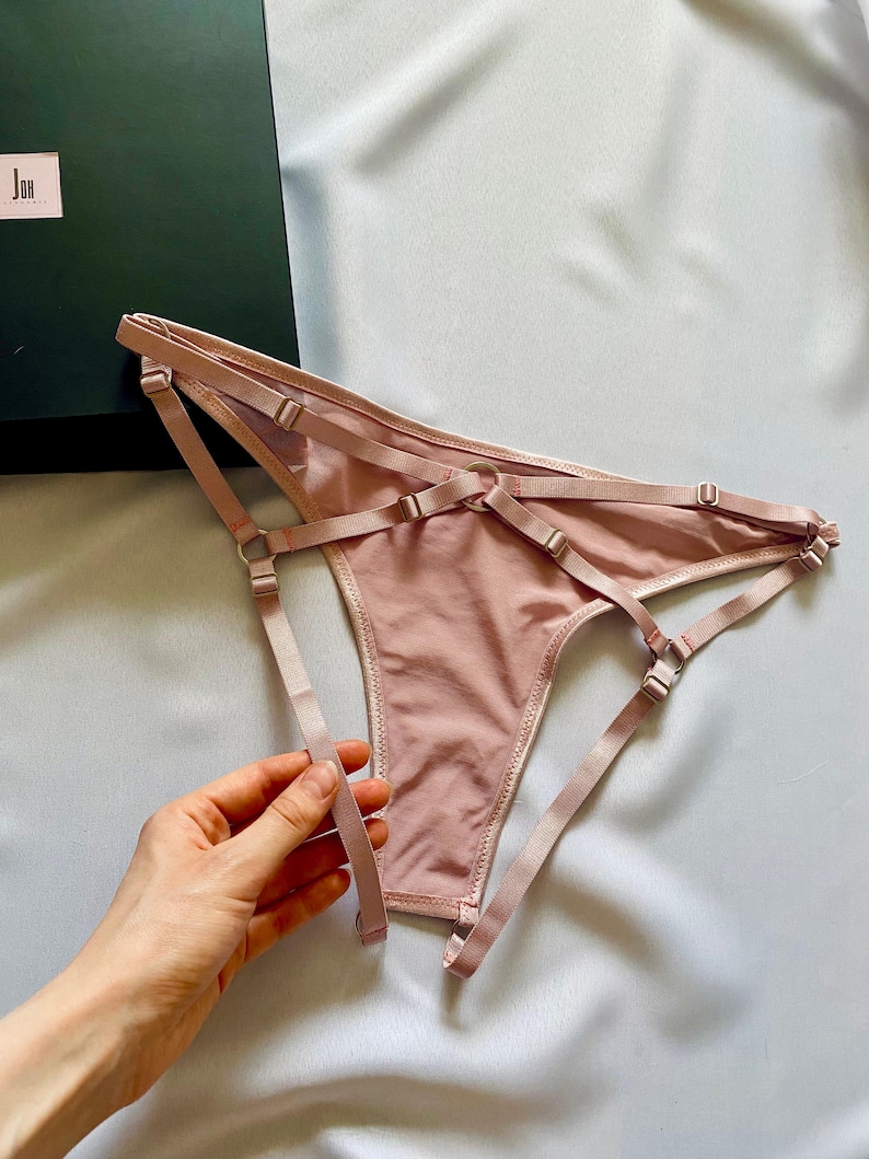 May include: Pale pink lingerie set with adjustable straps and delicate stitching. The set includes a thong-style bottom with multiple straps and metal hardware. A dark green box is in the background.