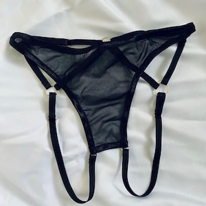 May include: Black lingerie set featuring a sheer thong design with multiple straps and gold-colored metal rings. The garment is made of a delicate, see-through fabric, and the straps create a unique, modern look. The lingerie is displayed on a white background.