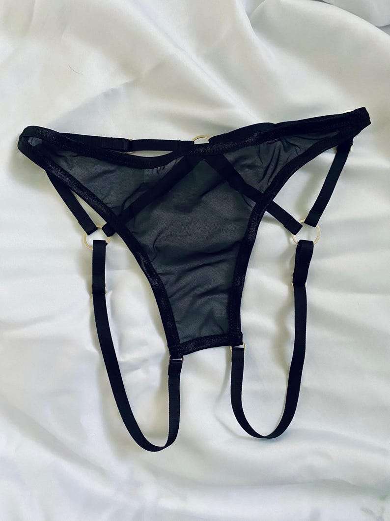 May include: Black lingerie set featuring a sheer thong design with multiple straps and gold-colored metal rings. The garment is made of a delicate, see-through fabric, and the straps create a unique, modern look. The lingerie is displayed on a white background.