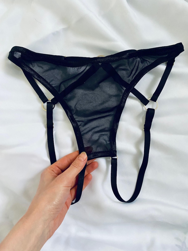 May include: Black lingerie set featuring a sheer thong design with a strappy, crisscross detail. The thong has a delicate, see-through mesh fabric and is accented with gold-colored metal rings. The lingerie is displayed on a white background.