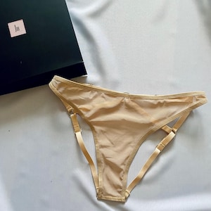May include: A beige thong-style underwear with adjustable straps and a matching waistband. The underwear is made of a sheer material. A black box with a small label is in the background.