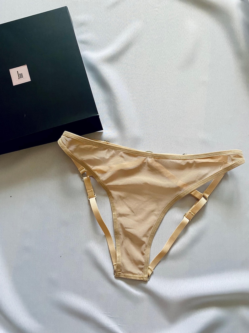 May include: A beige thong-style underwear with adjustable straps and a matching waistband. The underwear is made of a sheer material. A black box with a small label is in the background.