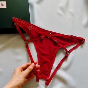 Puede incluir: Una tanga de color rojo brillante que presenta un complejo sistema de tiras elásticas finas conectadas por pequeños anillos metálicos dorados y deslizadores ajustables. La prenda tiene un diseño minimalista con múltiples aberturas.