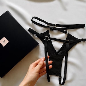 Puede incluir: Ropa interior negra con tiras que presenta un diseño complejo de bandas elásticas conectadas por anillos de metal plateado. La prenda incluye deslizadores ajustables en las correas y se presenta junto a una caja rectangular de color negro.