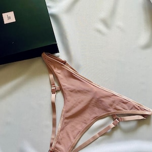 May include: A blush-colored lingerie set with adjustable straps and delicate stitching. The set is displayed on a light blue satin surface, with a dark green box in the background. The lingerie features a garter-style design.