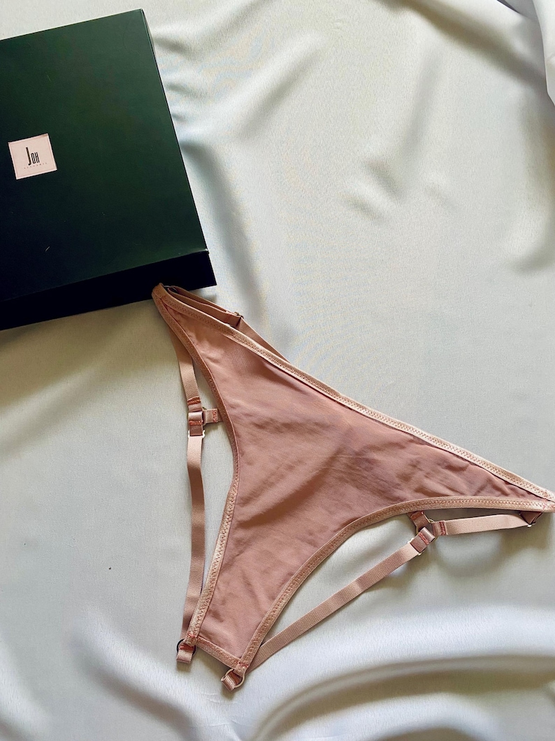 May include: A blush-colored lingerie set with adjustable straps and delicate stitching. The set is displayed on a light blue satin surface, with a dark green box in the background. The lingerie features a garter-style design.