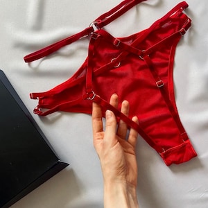 May include: A vibrant red thong featuring a complex system of adjustable elastic straps and small metal rings. The garment is constructed from a sheer, lightweight fabric and includes multiple sliders for fit customisation, presented alongside a black box.