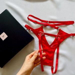 Puede incluir: Una tanga de tiras de color rojo brillante con múltiples bandas elásticas finas, anillos metálicos de tono plateado y deslizadores ajustables. La prenda se presenta junto a una caja rectangular negra con una pequeña etiqueta blanca con texto negro.