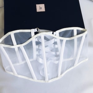 May include: White mesh corset with white satin ribbon lacing in the back. The corset has a white satin trim around the edges.