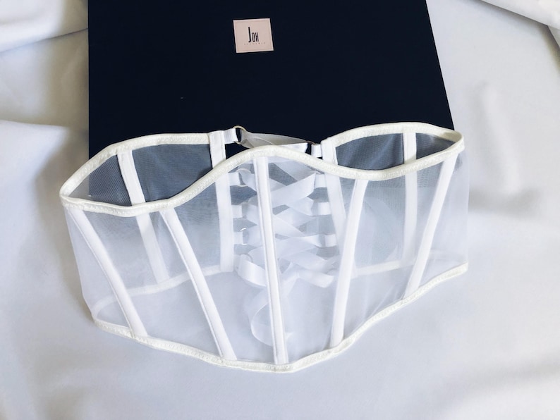 May include: White mesh corset with white satin ribbon lacing in the back. The corset has a white satin trim around the edges.