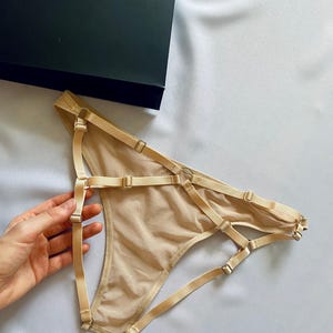 May include: Nude-colored lingerie with adjustable straps and metal hardware. The delicate underwear is displayed on a light-colored fabric, next to a dark green box with a white logo. The design features multiple straps and a sheer fabric.