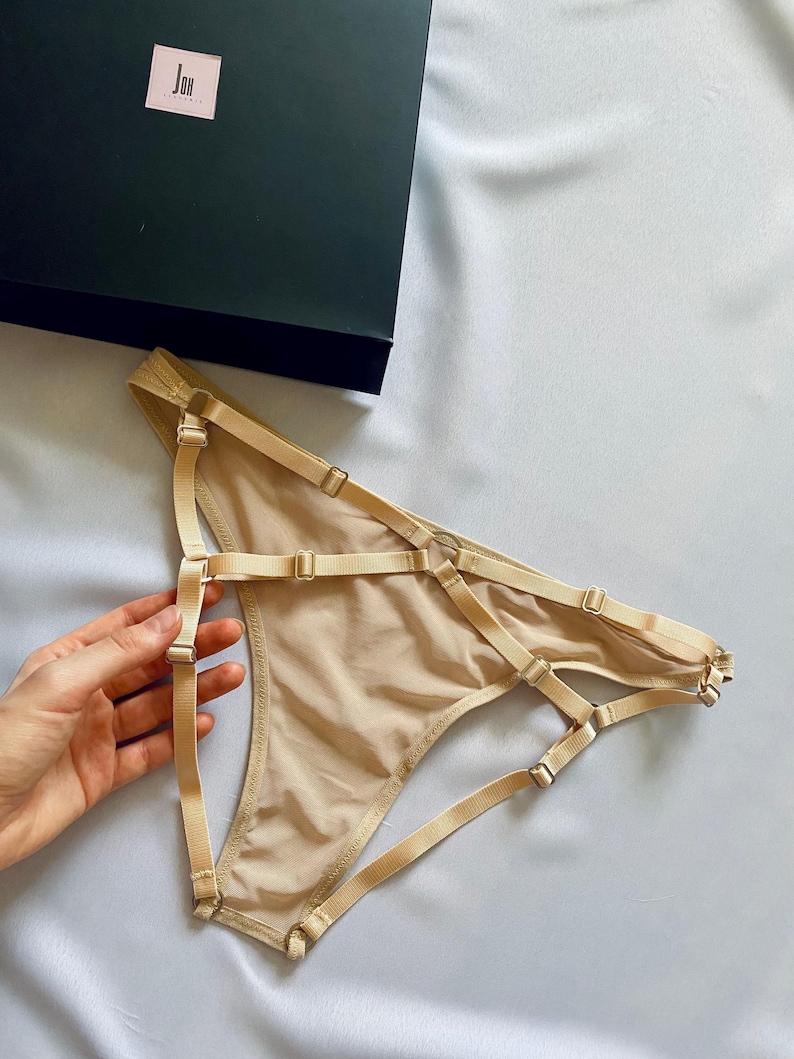May include: Nude-colored lingerie with adjustable straps and metal hardware. The delicate underwear is displayed on a light-colored fabric, next to a dark green box with a white logo. The design features multiple straps and a sheer fabric.