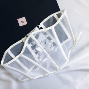 May include: White mesh corset with a lace-up front and a satin trim. The corset is on a white fabric background.