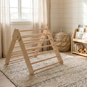 Peut inclure: Un triangle d'escalade en bois clair avec plusieurs barreaux, posé sur un tapis texturé. La pièce comprend une étagère avec des livres pour enfants, un panier tressé et un petit banc. Les murs sont blancs avec des panneaux horizontaux.